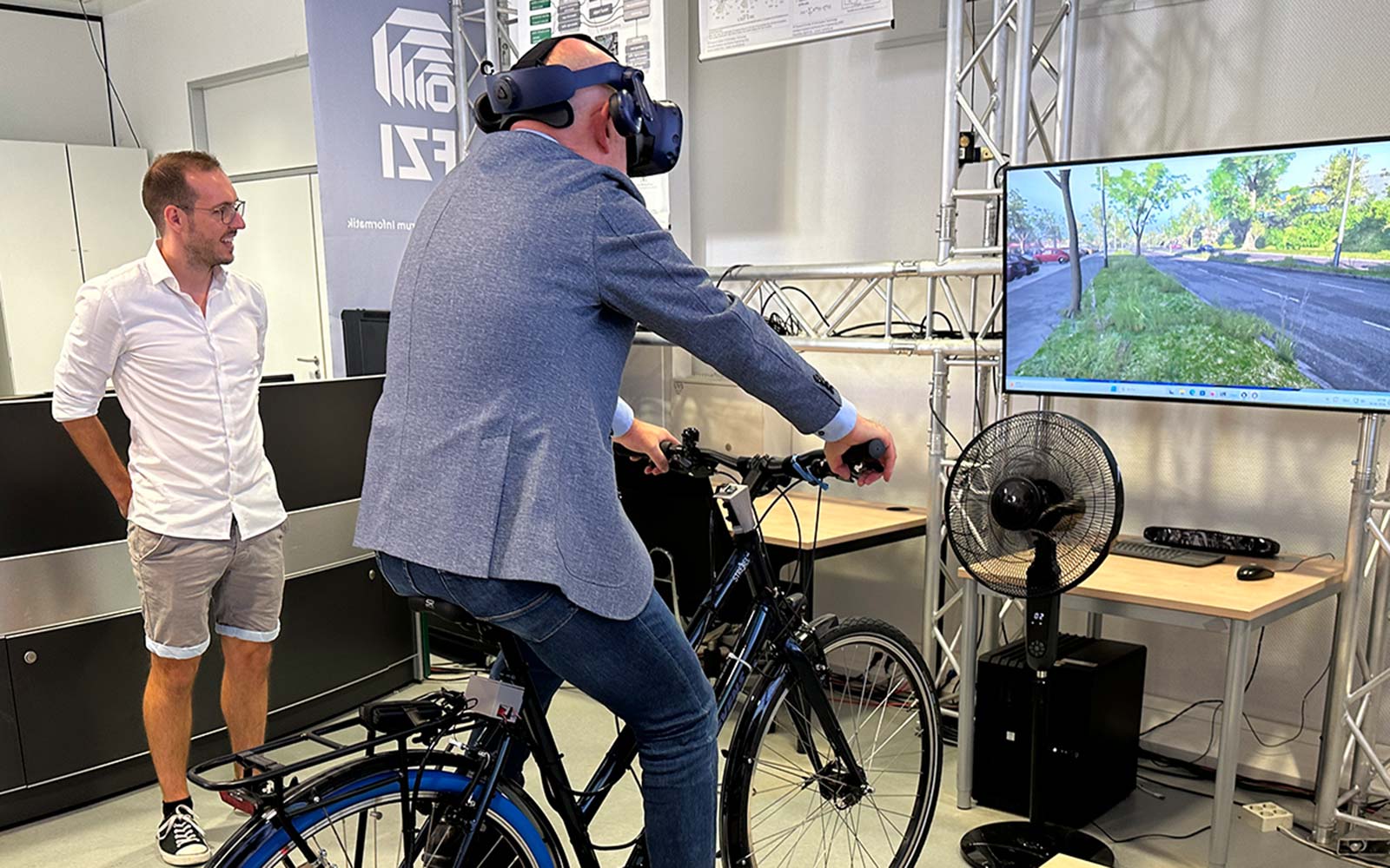 Verkehrsminister Winfried Hermann tritt beim FZI-Fahrradsimulator in Begleitung von FZI-Mitarbeiter Maximilian Zipfl in die Pedale